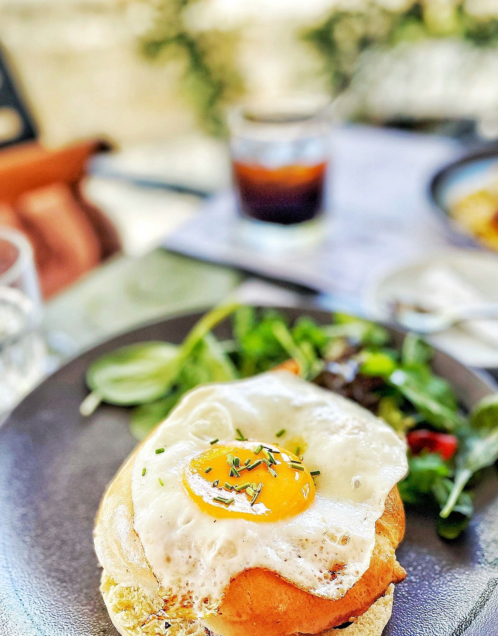A breakfast plate featuring a sunny-side-up egg over toast, served with a fresh green salad and a drink in the background.