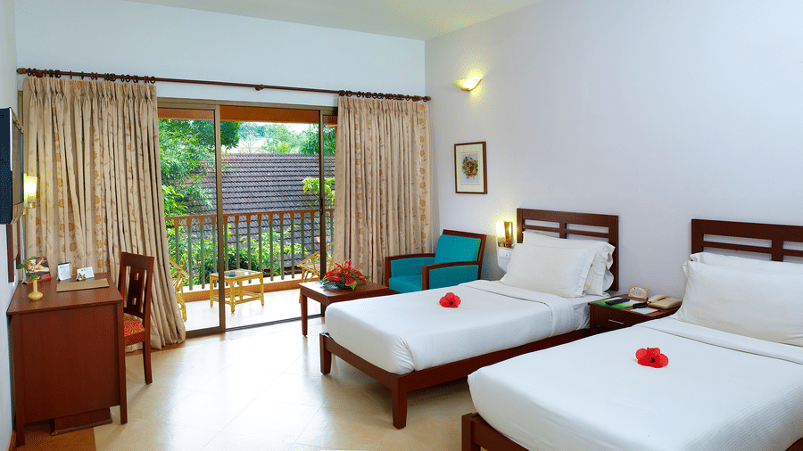 A bright room at Abad Green Forest, Thekkady with 2 single beds, a desk, and a balcony overlooking greenery.
