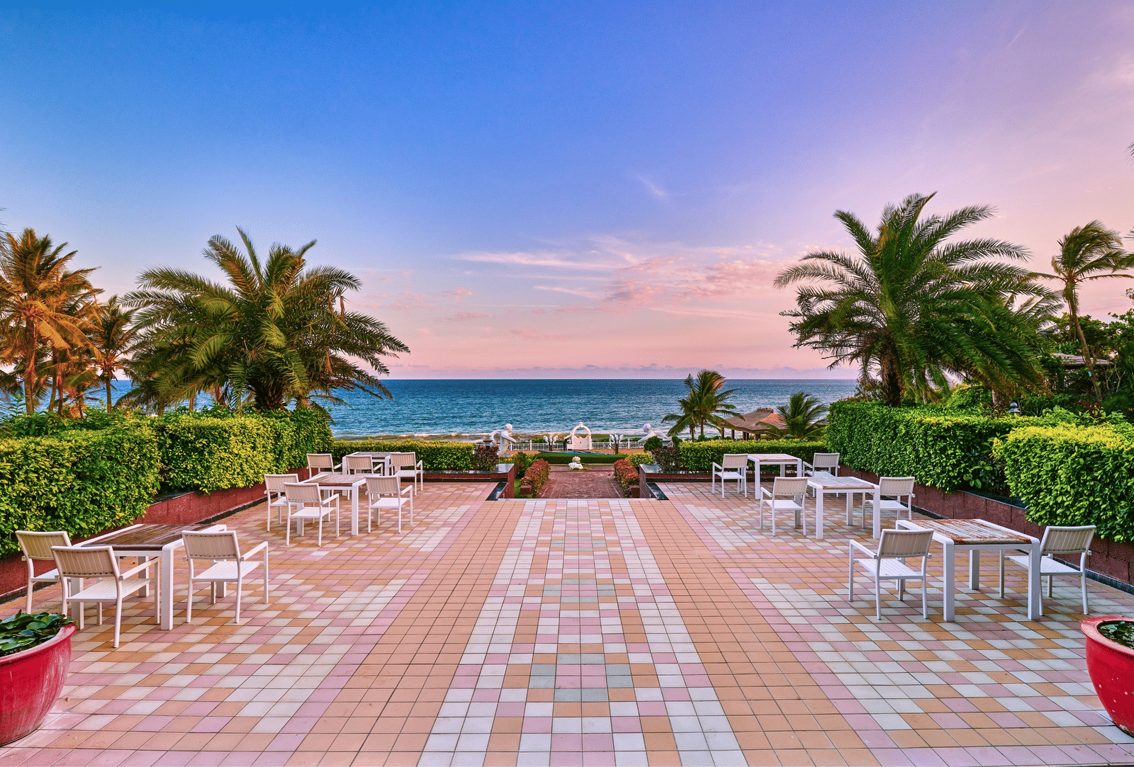 Open tiled terrace with white tables and chairs, clipped hedges and palm trees overlooking the sea at dawn at MAYFAIR Palm Beach Resort, Gopalpur-on-Sea.