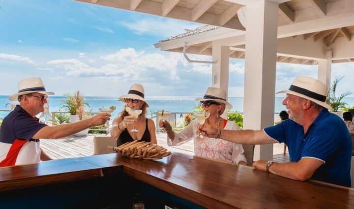 Four friends toast by the beachside bar, wearing sunhats and sharing laughter against a backdrop of blue skies and ocean views - Tamarind Hills Resort and Villas - Antigua