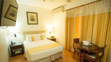 A bright bedroom with a bed, nightstands, and a dining table set near a window with yellow curtains | Economy Room | Altamont Court, Jamaica