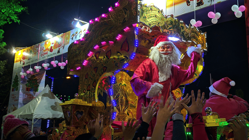 A lively outdoor festival or carnival area glows with bright lights and colourful decorations at night, populated by attendees.