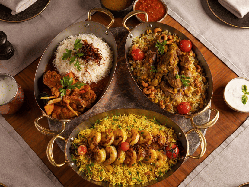 An overhead view of three oval serving dishes filled with food on a table. The dishes contain rice, meat, and shrimp, garnished with herbs and tomatoes. Two small bowls of sauces and a glass of yogurt drink are also visible.
