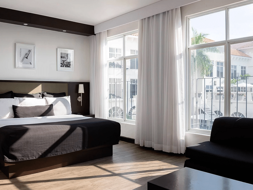 An image of a Jamaican hotel room with floor-to-wall windows, a large bed and a couch - S Hotel Kingston, Jamaica