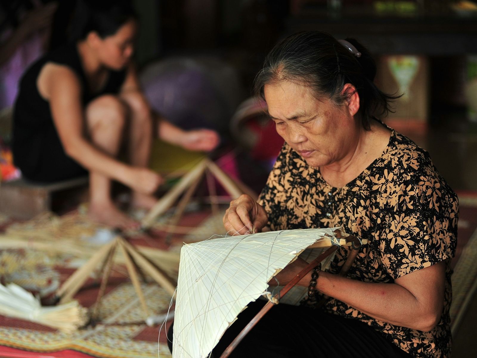 A close up of an old woman hand weaving a conical hat with another woman doing the same in the background.
