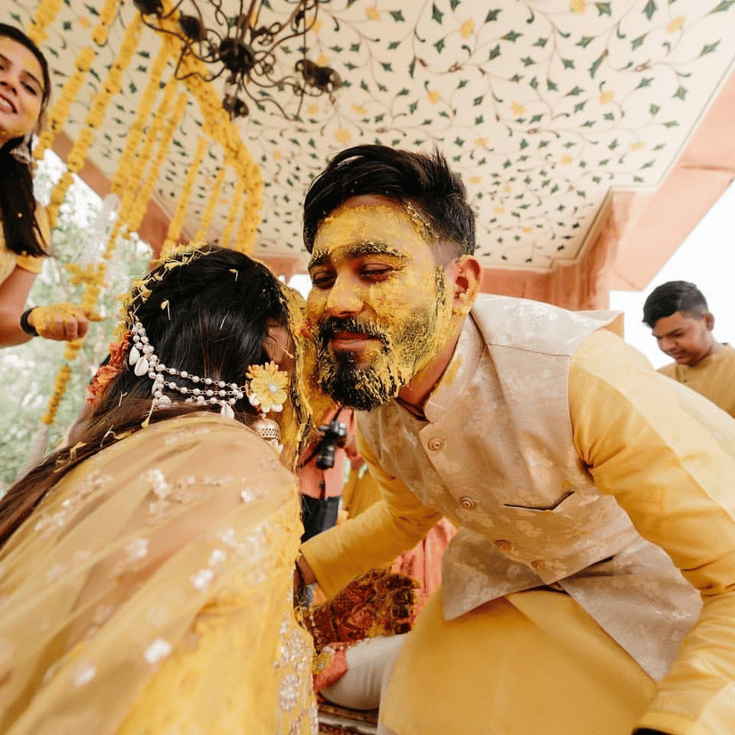 A groom with turmeric-covered face leans in playfully towards his bride during a joyful haldi ceremony, surrounded by loved ones.- Umaid Palace - A Lakeside Heritage Palace Resort