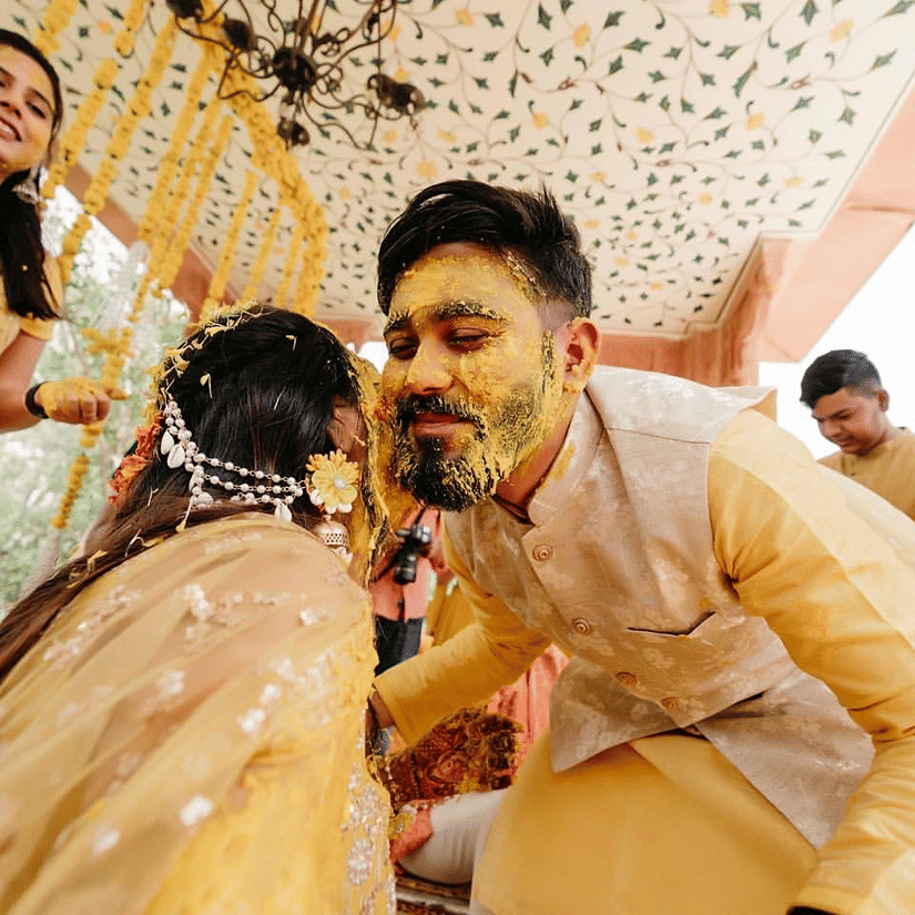 A groom with turmeric-covered face leans in playfully towards his bride during a joyful haldi ceremony, surrounded by loved ones at Umaid Palace, one of the royal wedding venues in Jaipur.