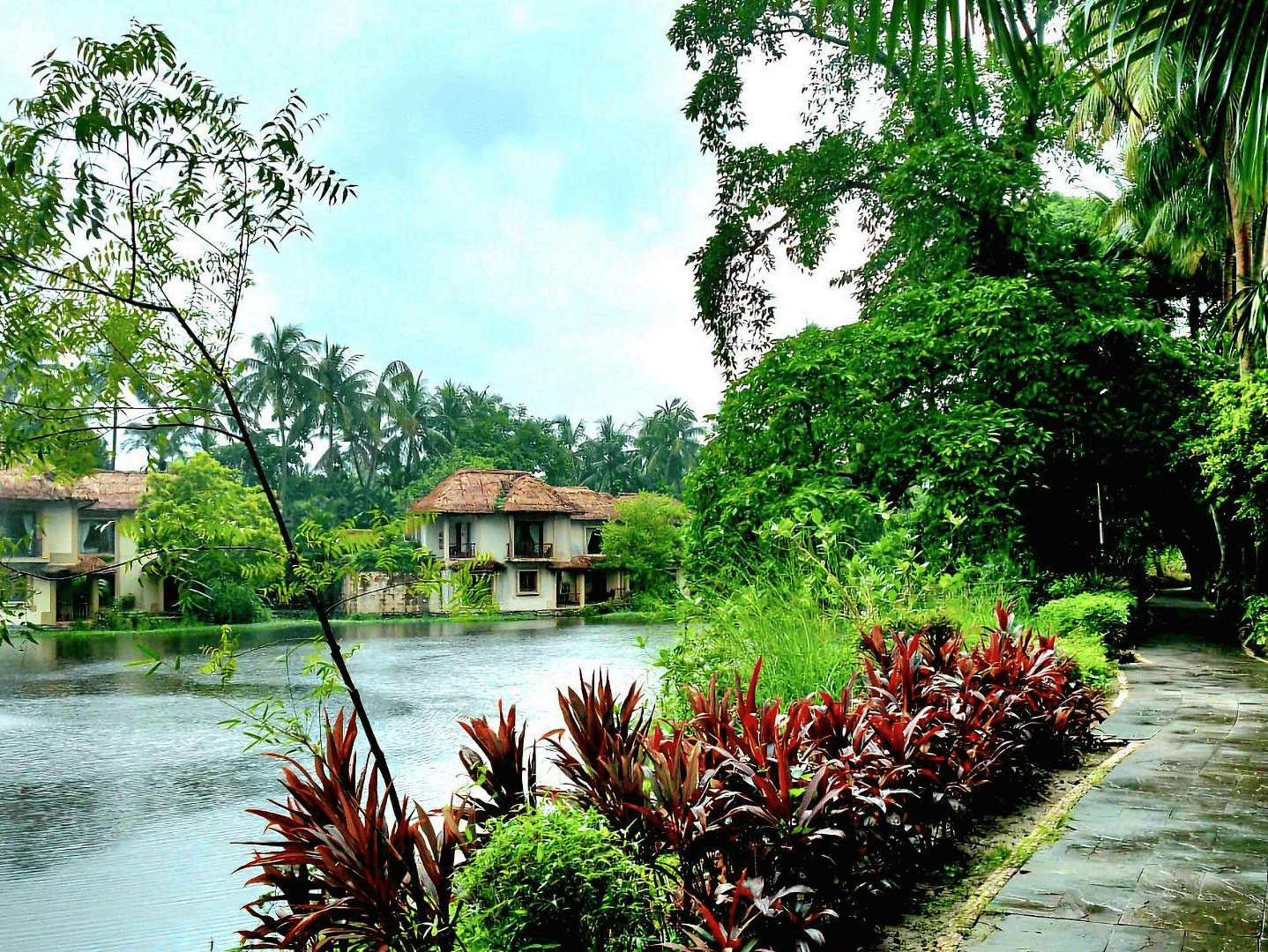 A serene waterside walkway with red and green plants and a fountain, featuring resort buildings in the background at the Vedic Village Spa Resort.