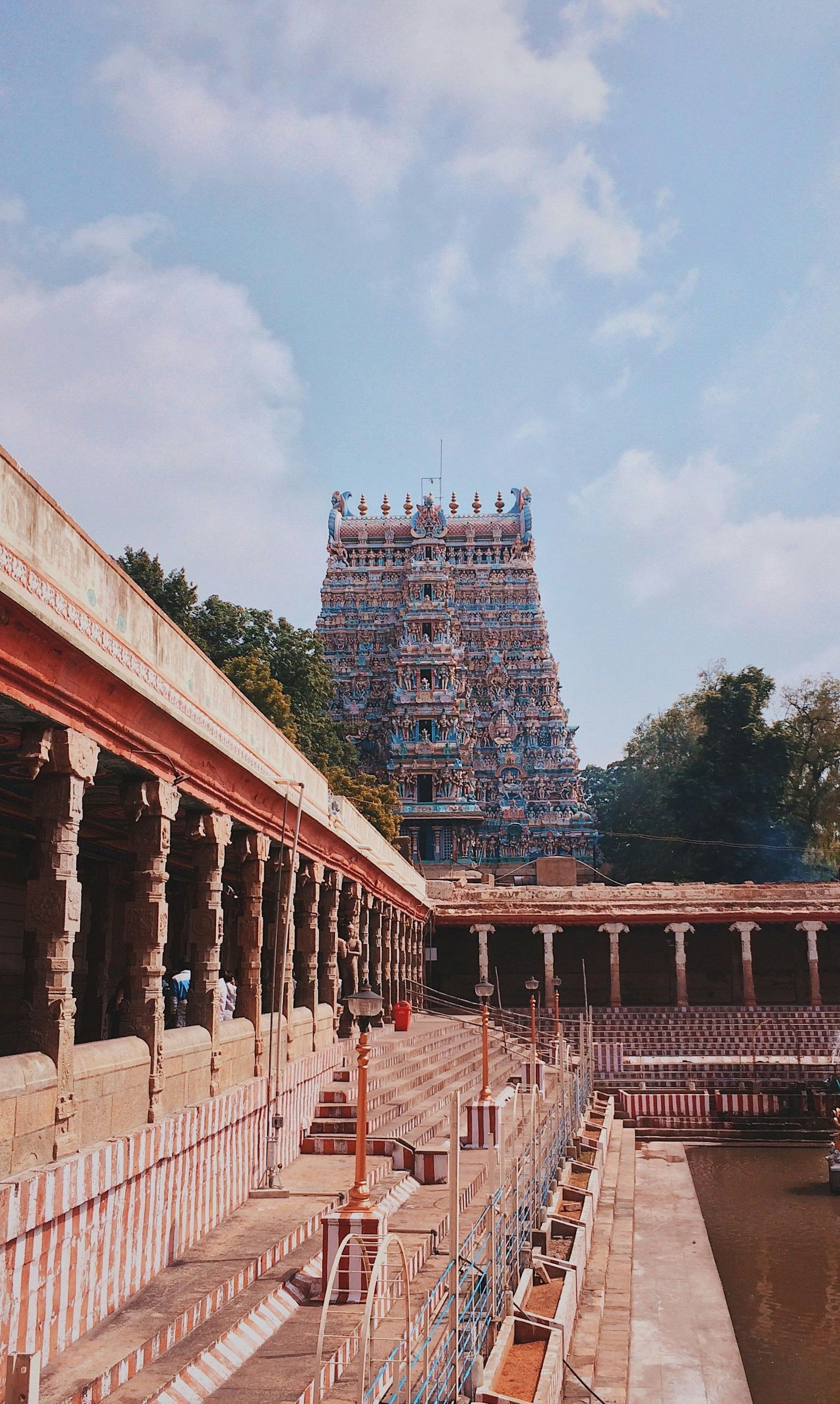 An image of a temple pond with steps and pillars, featuring a five-tiered gopuram in Dravidian architectural style in the back