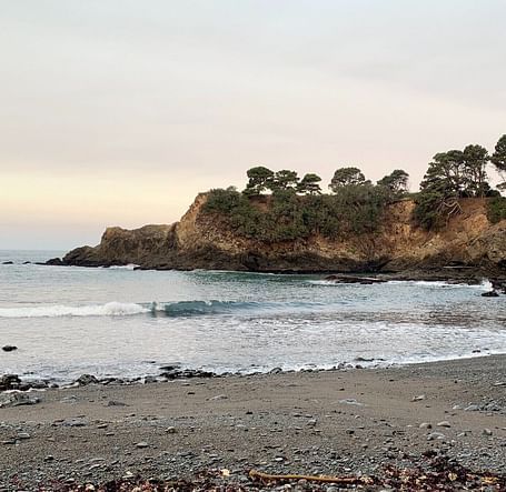 A secluded beach with dark sand and driftwood logs sits nestled between rocky cliffs under a cloudy sky.
