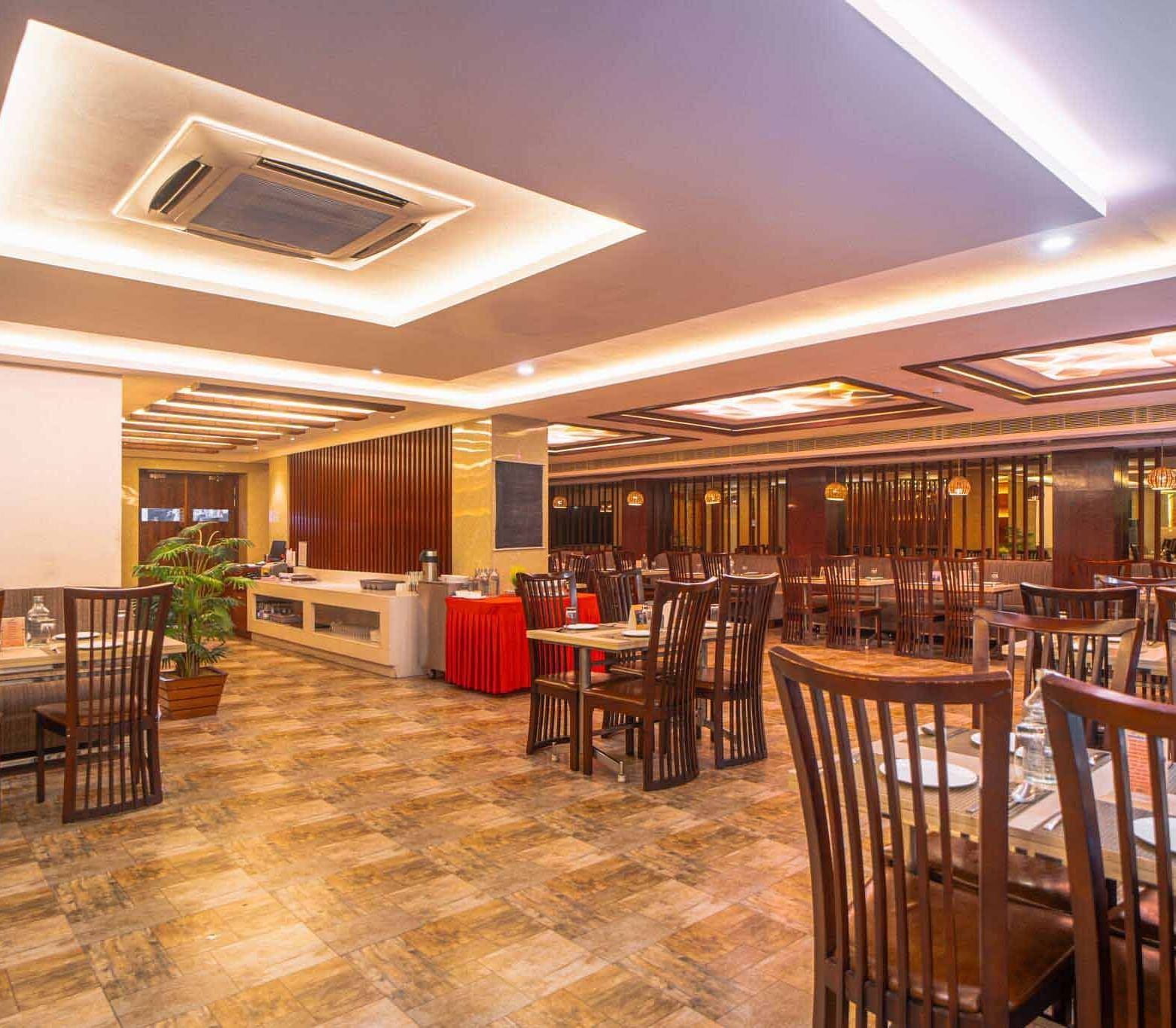 A wide shot of a spacious dining area with wooden tables and chairs, illuminated by warm ceiling lights at Mansha Hotel Regency.