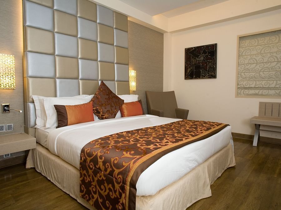 A hotel room featuring a bed with a headboard, a side table with a lamp, and a window with blinds- The Sonnet, Kolkata.