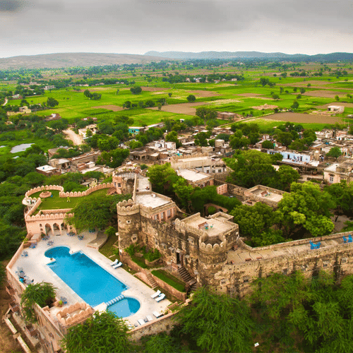 Aerial drone shot of Hill Fort Kesroli 14th Century Alwar where swimming pool and multiple structures are surrounded by lush green landscape and cloudy blue sky in the background.