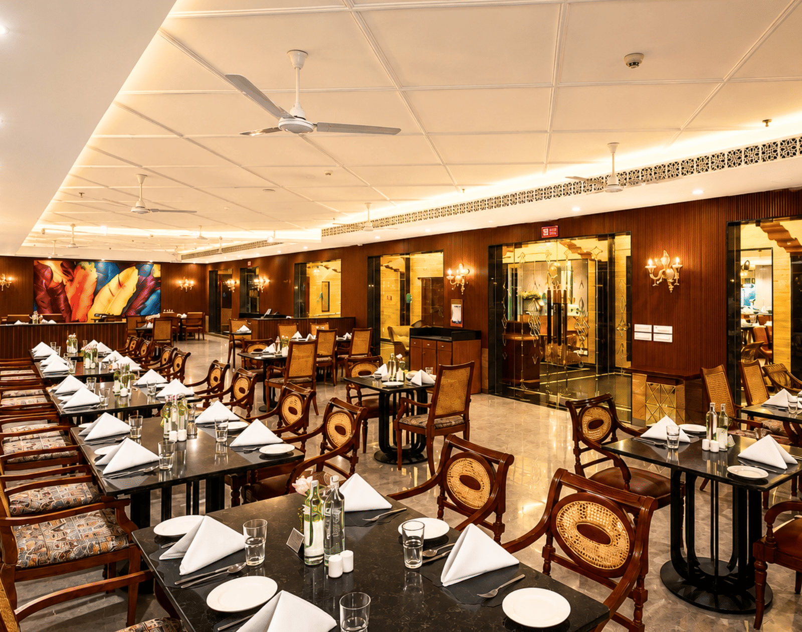 Indoor 1914 restaurant with marble-topped tables, cane-back chairs, wood-panelled walls and feather wall art at MAYFAIR Palm Beach Resort, Gopalpur-on-Sea.