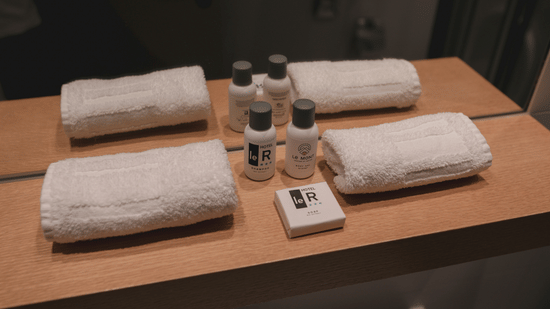 A variety of small hotel toiletries including shampoo, body wash, soap, and rolled white towels are arranged on a wooden shelf.