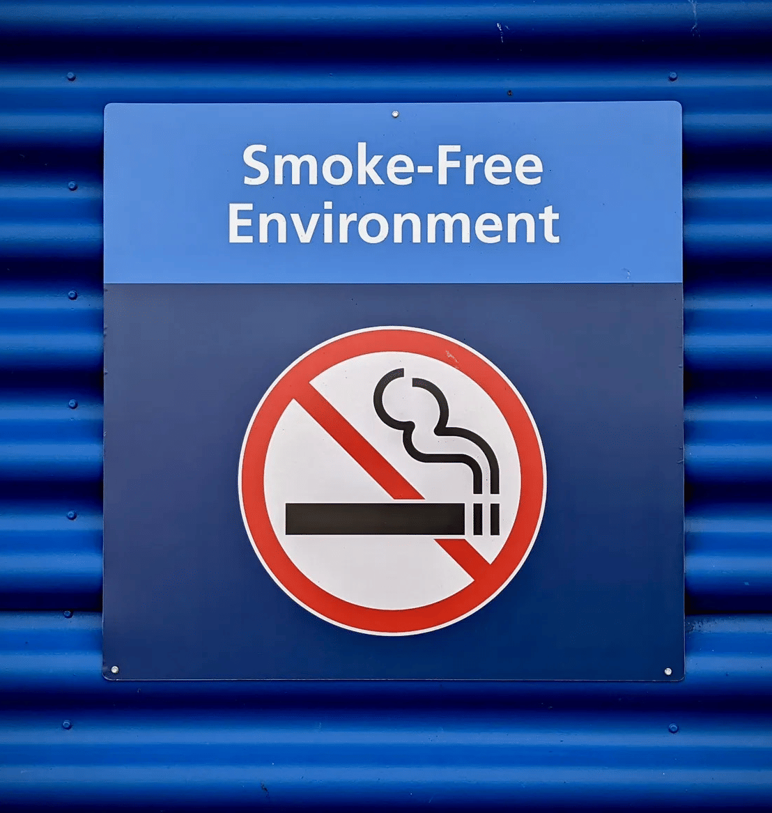A blue sign reading 'Smoke-Free Environment' with a no-smoking symbol is attached to a corrugated wall.