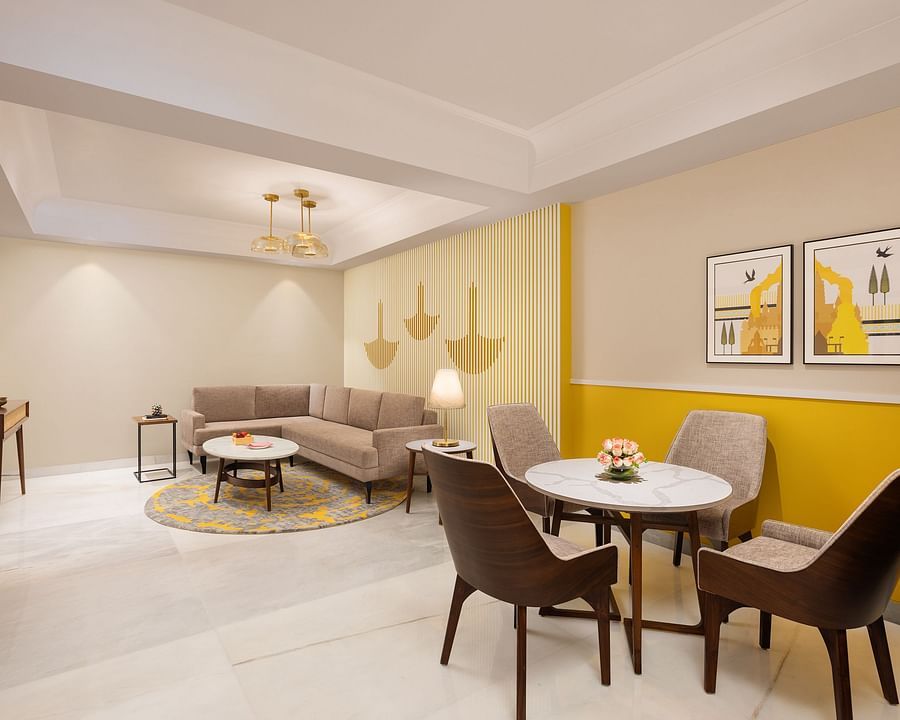 A bright living and dining area with a beige sectional sofa, a small round table on a yellow-patterned rug, a dining table with four chairs, a yellow and white striped accent wall with abstract art, and a television on a wooden stand - Aloha On the Ganges.