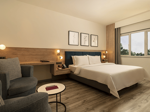 A warmly lit Premium Room features a double bed with white linens, two armchairs, a small round table, and framed pictures above the headboard at The Residency Karur.