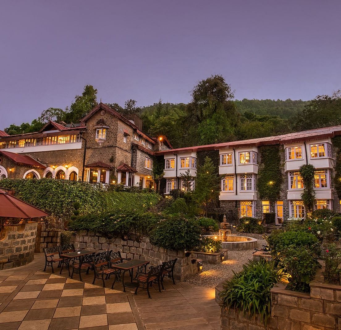 A scenic view of a the naini retreat, nainital, The resort features multiple buildings with stone exteriors and white trim. The surrounding landscape includes mountains, trees, and a winding pathway.