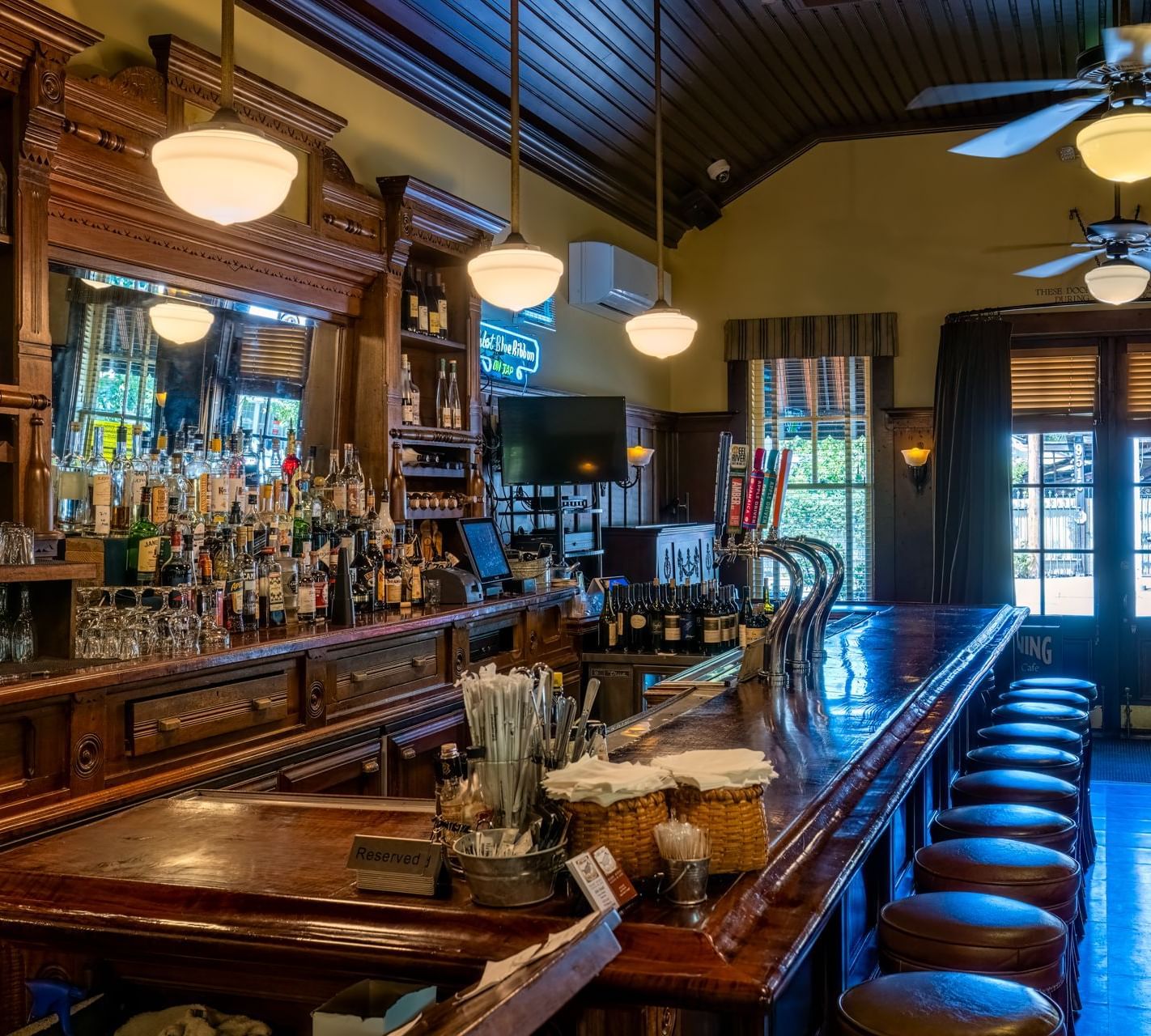 The rustic interior of a lively bar with a long wooden counter, bar stools, and various beverages at The Tallman Hotel.