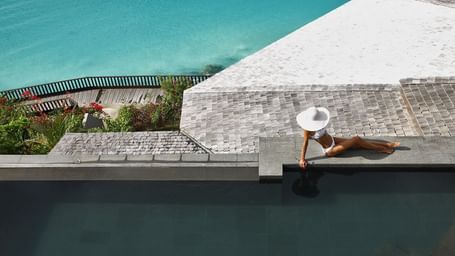 An overhead view of a woman in a wide-brimmed hat lounging by a dark infinity pool overlooking a vibrant turquoise ocean.