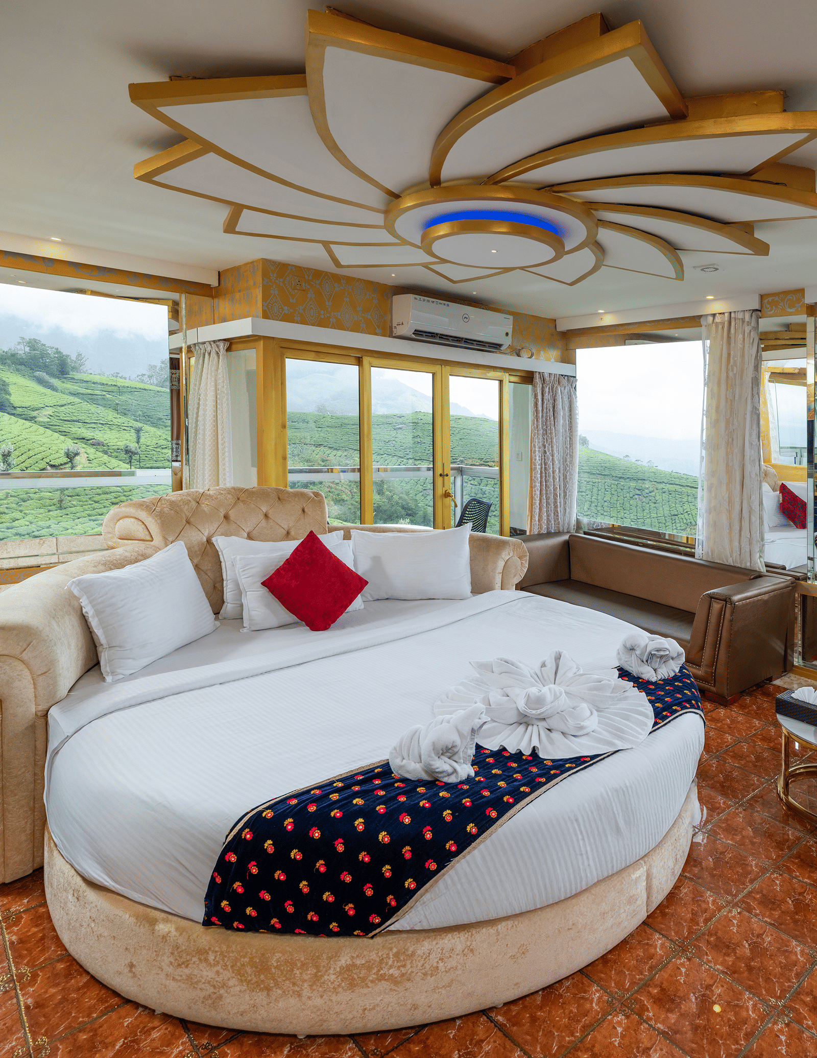An interior view of the Jacuzzi 180-degree Suite at Parakkat Nature Hotel and Resorts, Munnar, featuring a large round bed, plush decorative pillows, and elaborate gold-toned ceiling accents.