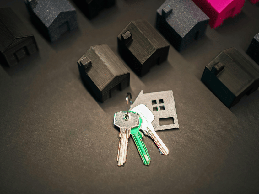 House-shaped models arranged on a dark surface with a set of keys and a small house keychain in the foreground, symbolising home ownership or real estate.