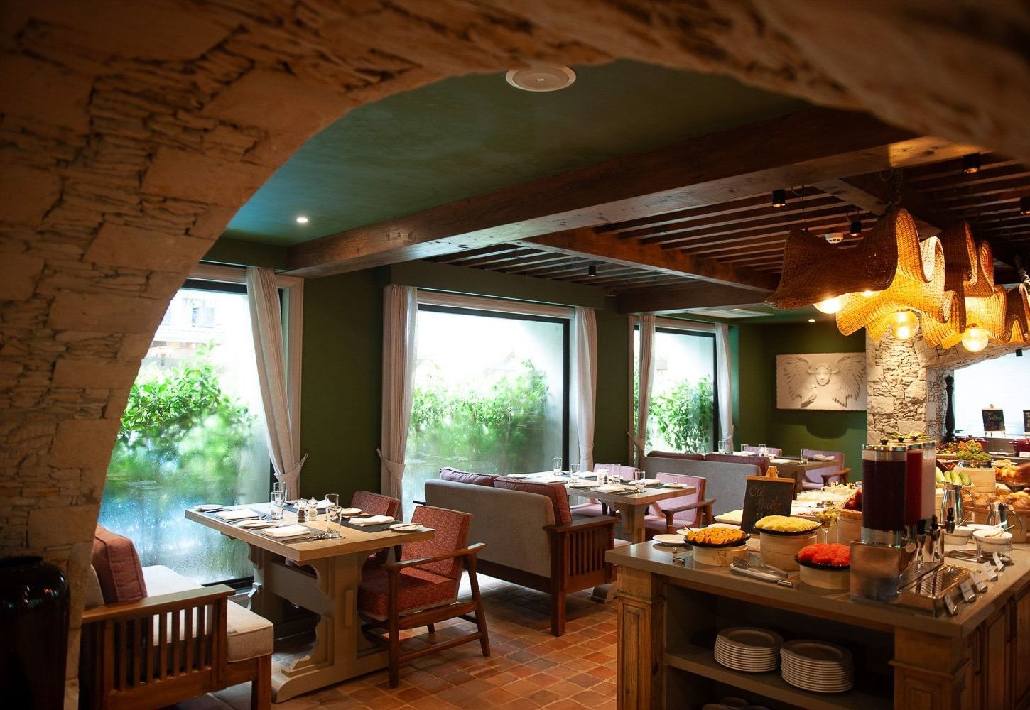 A wide-angle view of the dining area at the Mila Restaurant at The Evren, Vagator, from the corner of the stone archway featuring wooden tables, comfortable chairs and banquettes, and a buffet station with a variety of food.