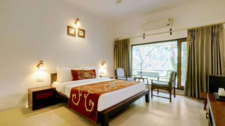 A bedroom featuring a wooden bed, 2 small bedside cabinets, 2 chairs facing a small table with a vase on it, and glass doors offering a view of the attached balcony and trees outside | Summit by the Ganges Beach Resort & Spa, Rishikesh 