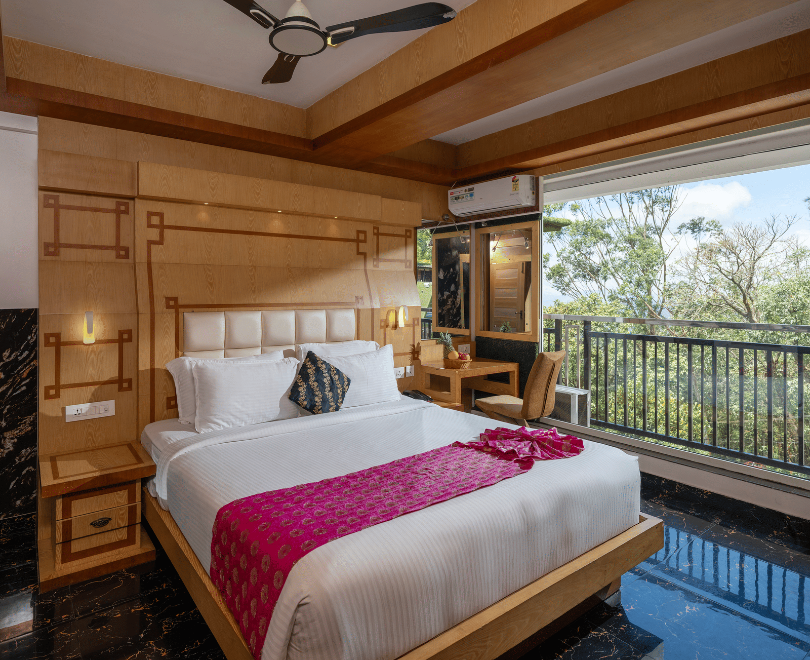 The interior of Superior Room at Parakkat Nature Hotel and Resorts, Munnar featuring a large bed, wooden panelling on the walls and ceiling, a ceiling fan, and a large window overlooking greenery.