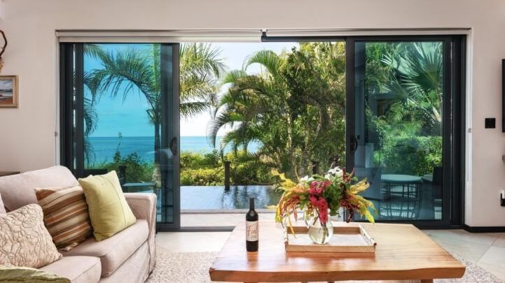 A living room with a couch, coffee table with wine and flowers, and large glass doors opening to an ocean view.