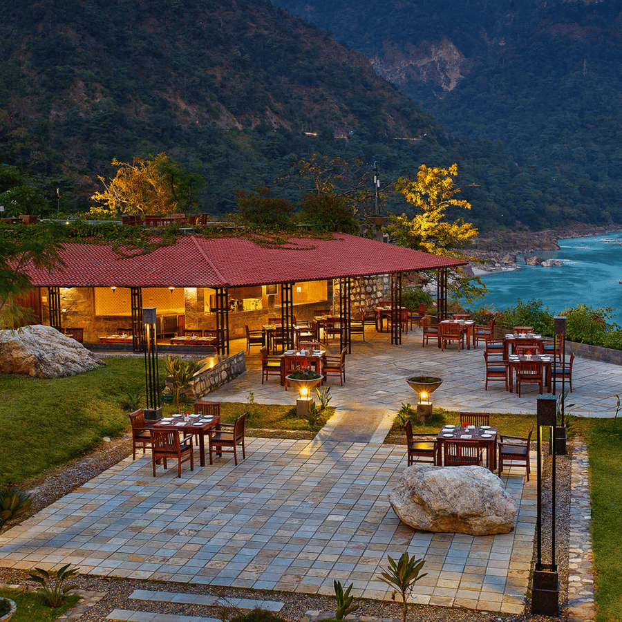 Outdoor dining area by a river, with tables set on a stone patio. A red-roofed pavilion is surrounded by lush greenery and mountains, conveying tranquility - Aloha On the Ganges