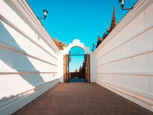 A long, open-air walkway with high white walls leading toward a decorative archway and blue sky.