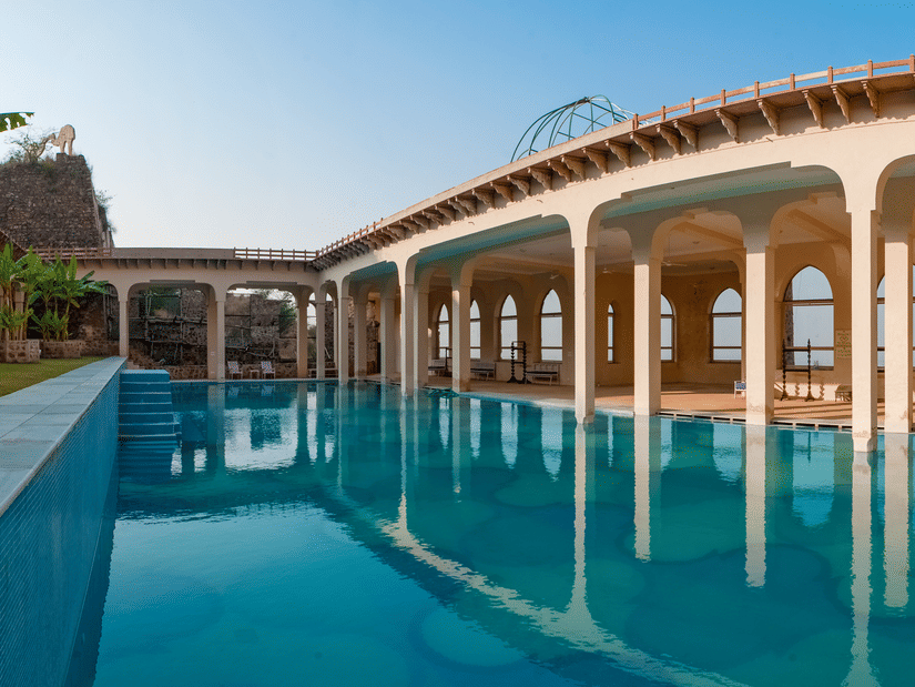 A long blue swimming pool reflects the arched architecture of Tijara Fort-Palace under a clear sky in Alwar.