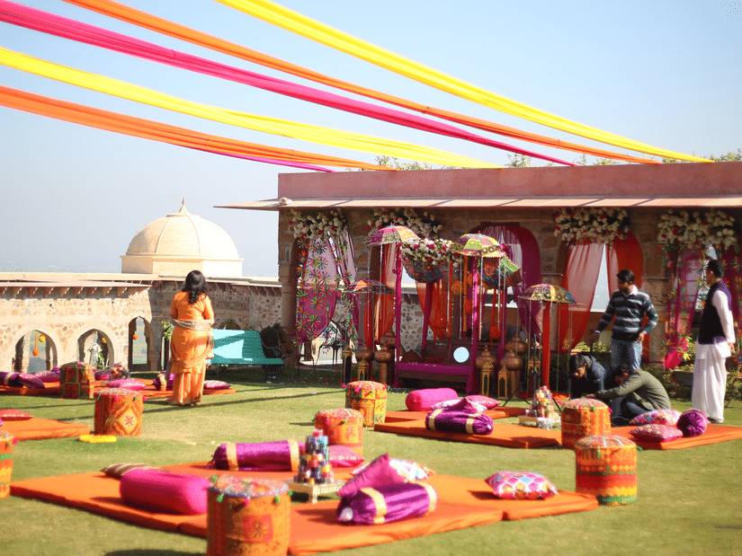 A vibrant outdoor event is set up on a green lawn at Tijara Fort-Palace - 19th Century, Alwar with clear blue sky in the background.