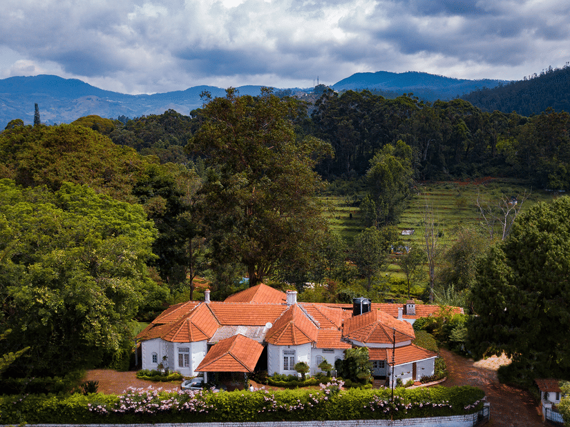 An aerial view of the hotel with trees surrounding it and a tea plantation in the background - Wallwood Garden - 19th Century, Coonoor