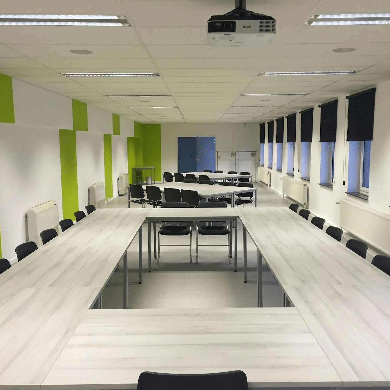 Image of a meeting room with tables and chairs and windows on the side. 