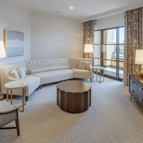 Warmly lit living room of Lakeview King Suite at YO1 Longevity & Health Resorts featuring a sofa by a window with coffee table facing the TV on the wooden counter and a couch next to the lamp near the side table of sofa