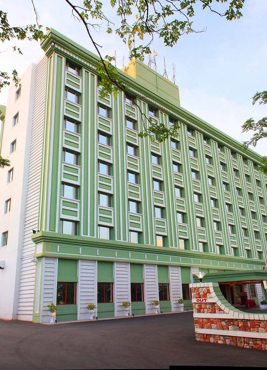 A multi-story green hotel building flanked by trees at Tara, Comfort Hotel in Hyderabad - Ramoji Film City.