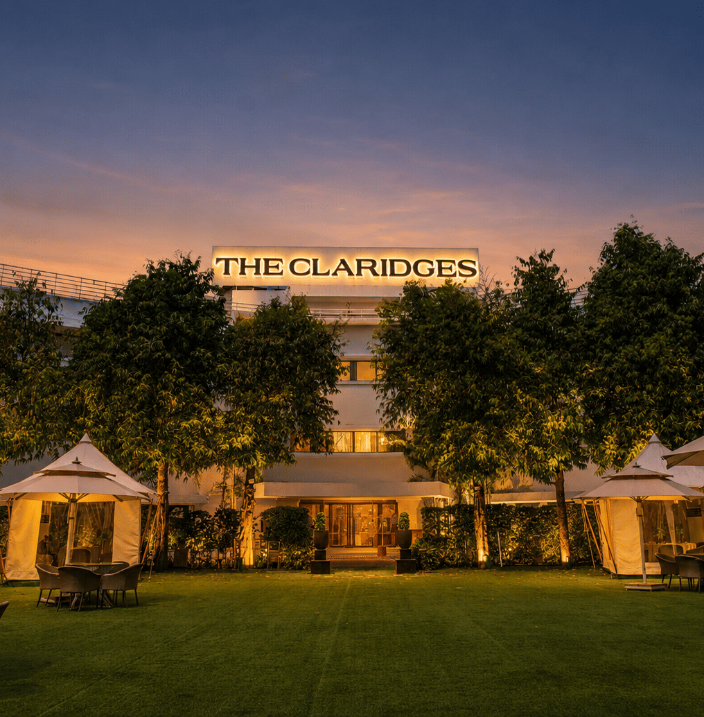 Evening view of a green hotel lawn featuring warm-lit white canopies and outdoor dining tables in front of an illuminated building at dusk - The Claridges New Delhi
