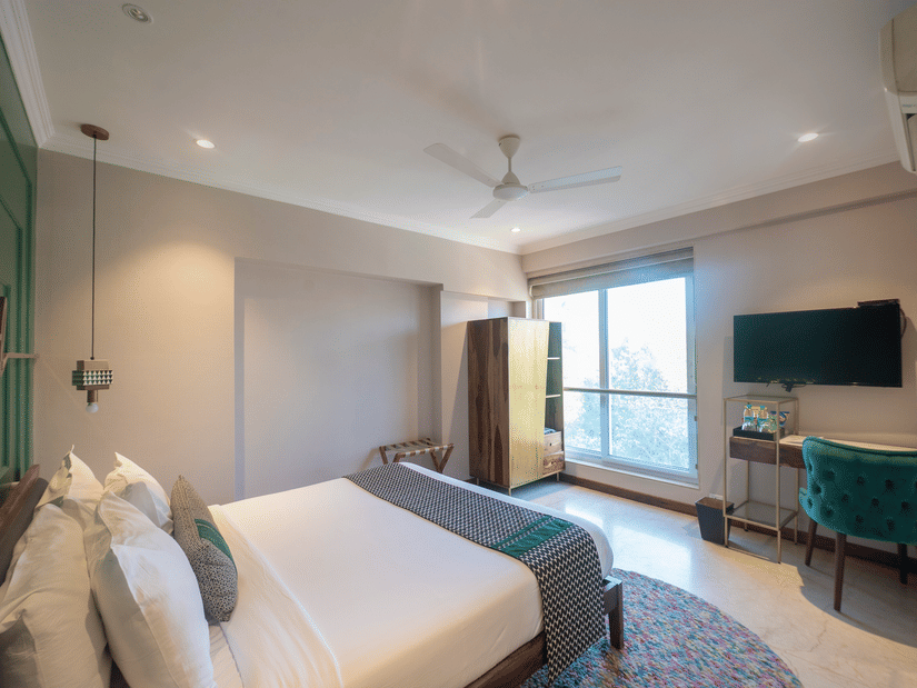 A bedroom with a neatly made bed, pendant lighting, ceiling fan, wardrobe, work desk with chair, wall-mounted TV, and a large window letting in natural light - Theory9 - Premium Service Apartments, Bandra