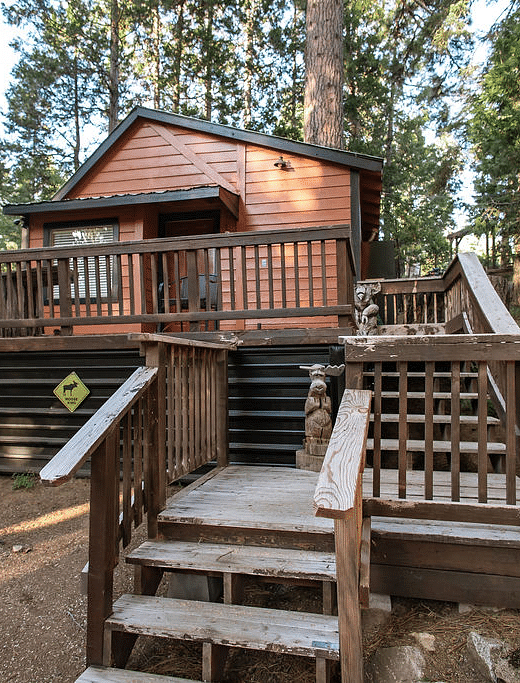 A Cabin at Shaver Lake Village Hotel with a wooden deck, stairs, and a grill, surrounded by tall pine trees.