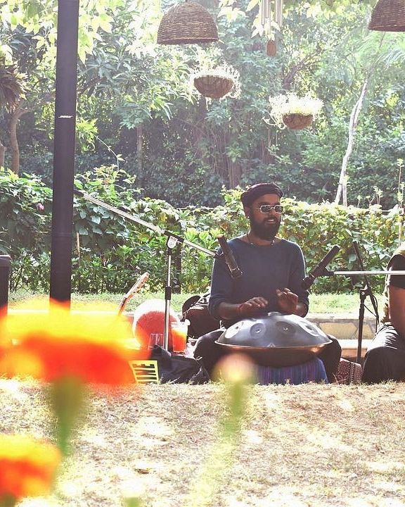 Two people sitting under an outdoor portico, surrounded by lush trees and plants, playing traditional instruments with audio equipment at Karma Chalets.