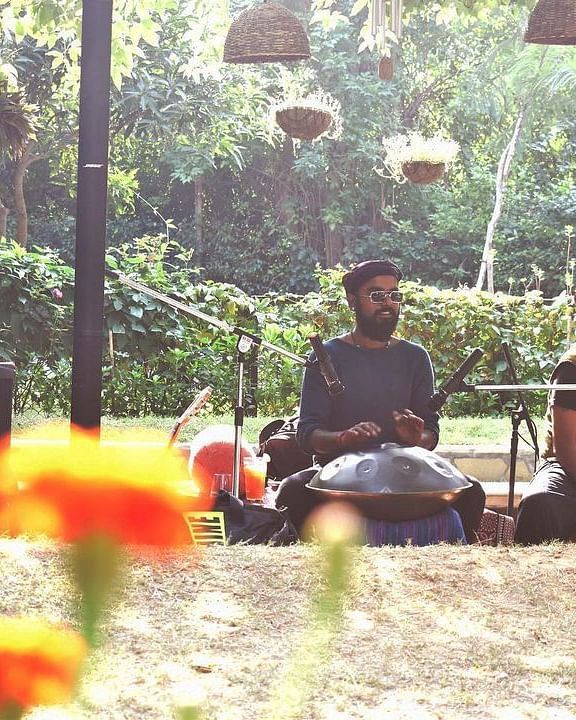 Two people sitting under an outdoor portico, surrounded by lush trees and plants, playing traditional instruments with audio equipment at Karma Chalets.