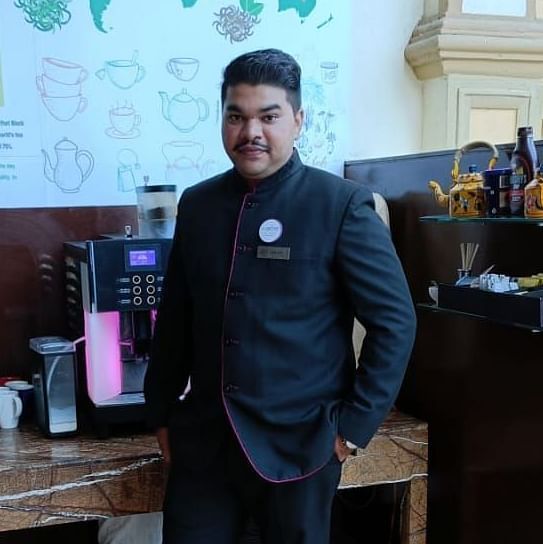 Hotel staff member in a formal uniform standing near a wooden beverage station with coffee and tea supplies, set against a wall featuring a world map poster.