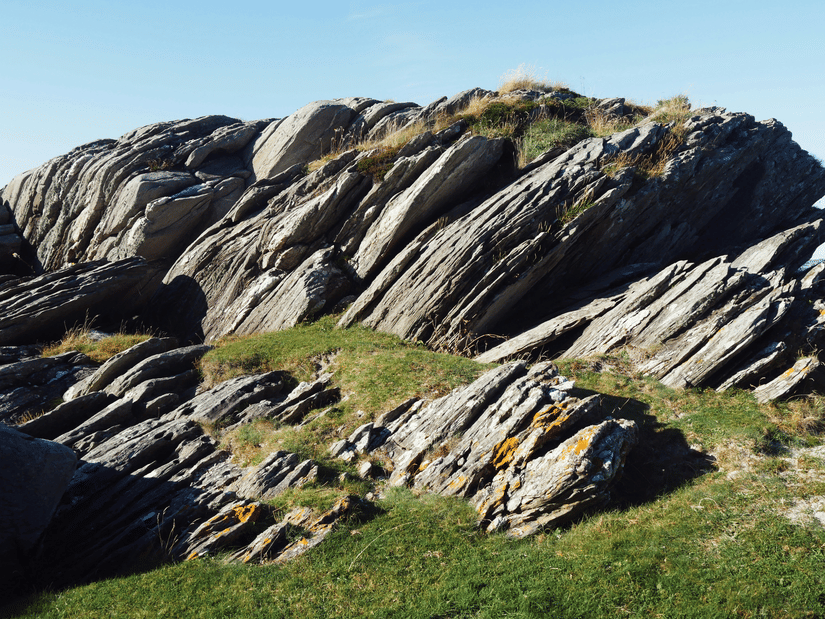 A close-up of a striking geological rock formation showing deeply stratified, angled grey layers with patches of green grass and lichen.