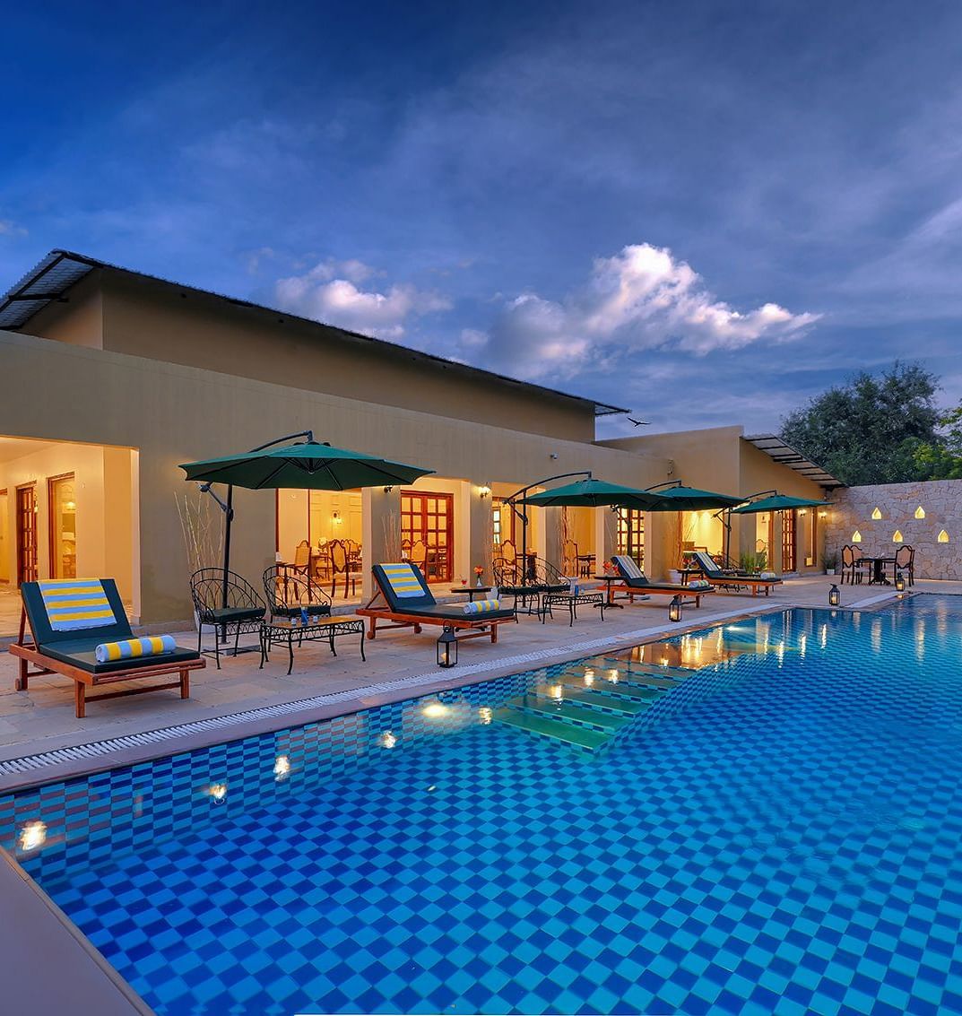 A swimming pool with checkered tiles, lounge chairs, and umbrellas in front of a building at dusk - SinghVilas Ranthambore