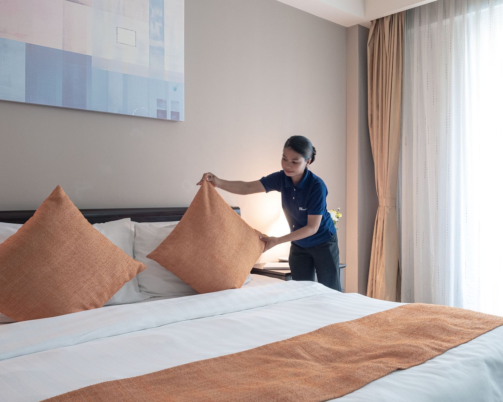 A staff member making a bed, adjusting pillows on a large white bed with orange decorative pillows and a matching bed runner | Central Mansions, Phnom Penh