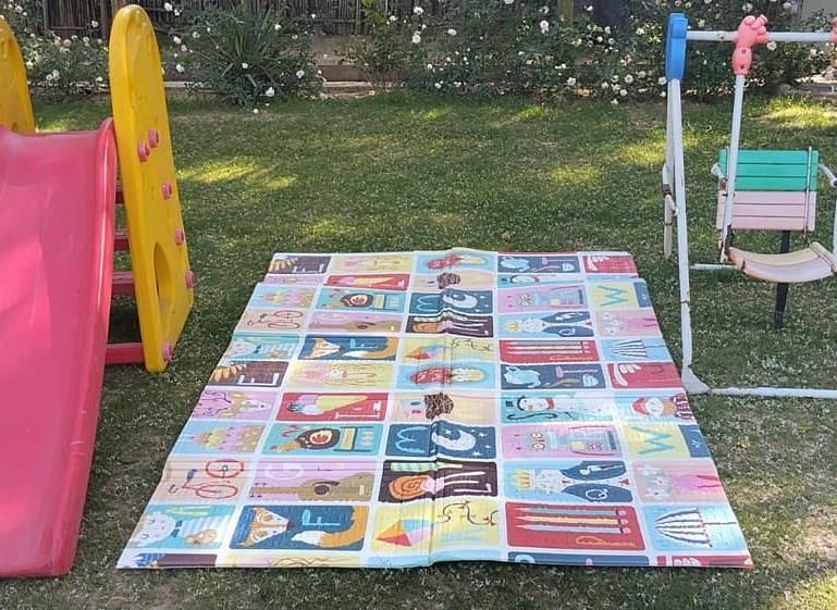 An outdoor play area featuring a small slide, a swing set, and a colourful play mat spread on the grass.