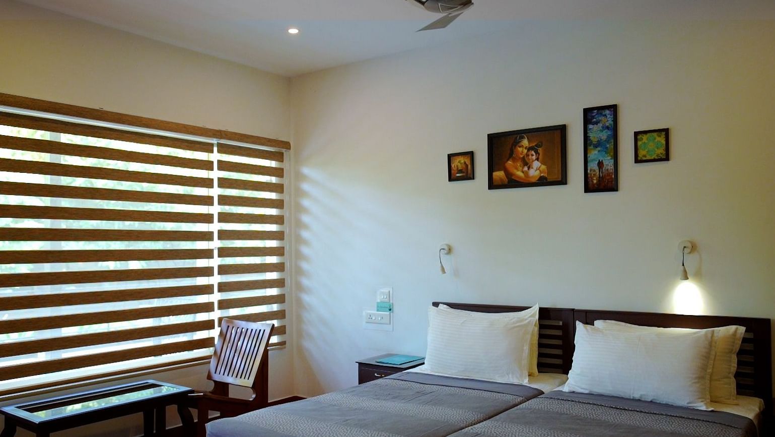 A bedroom with two twin beds pushed together, white pillows, a dark wooden headboard, framed artwork on the wall, and striped zebra blinds on the window.